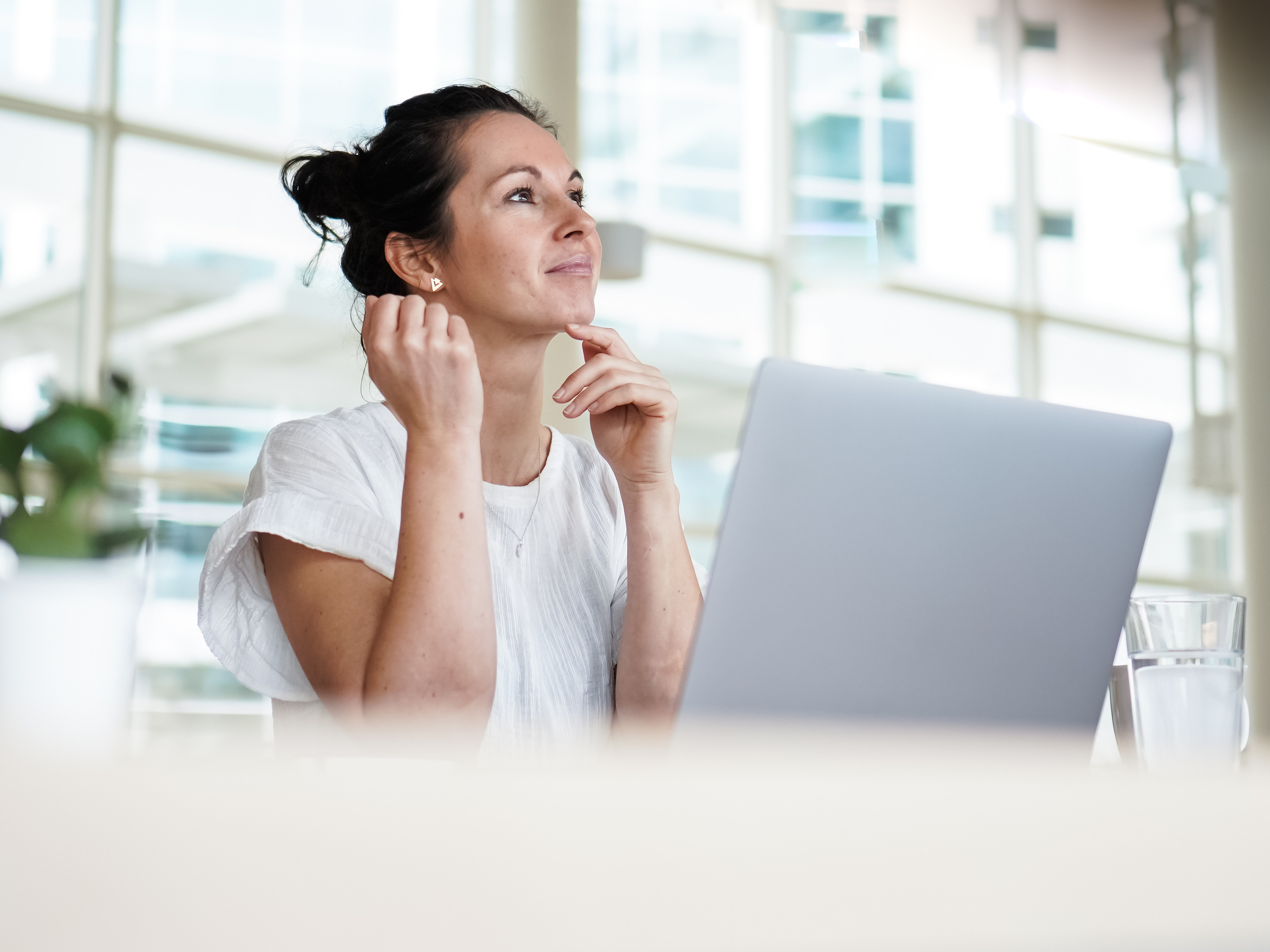 Eine Frau sitzt mit einem Laptop an einem Schreibtisch, lächelt nachdenklich und blickt nach oben. Sie trägt ein weißes Oberteil. In der Nähe stehen ein Glas Wasser und eine kleine Topfpflanze in einem hellen, modernen Büro.
