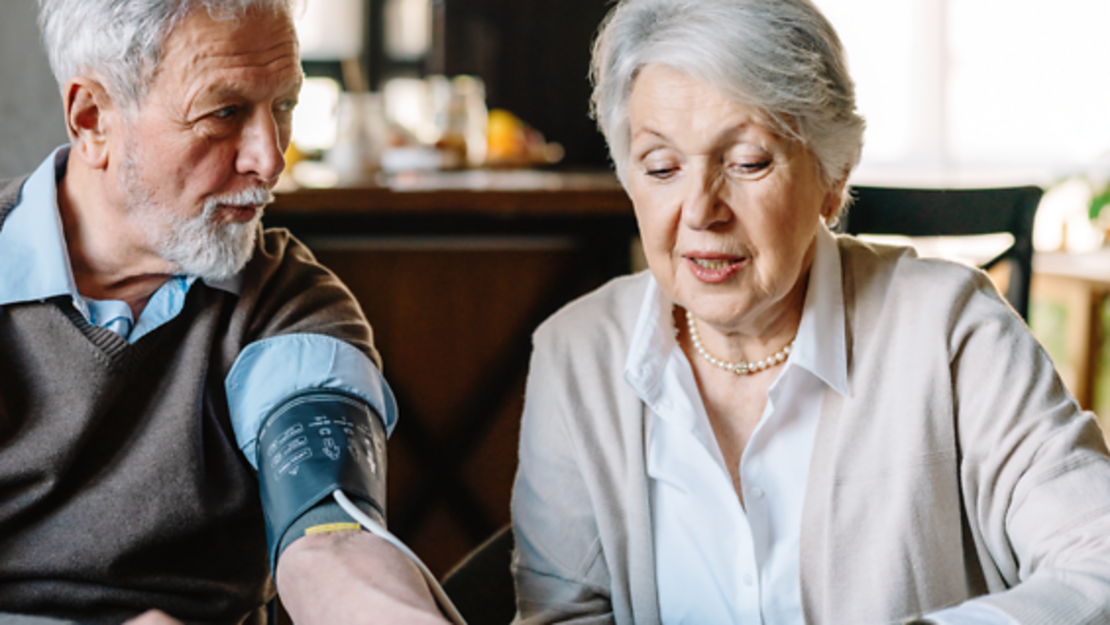 Ein älterer Mann lässt seinen Blutdruck von einer älteren Frau zu Hause messen. Das Bild zeigt ein blaues Banner mit deutschem Text über Notfallmanagement bei Herzinsuffizienz und das Erkennen kritischer Situationen.