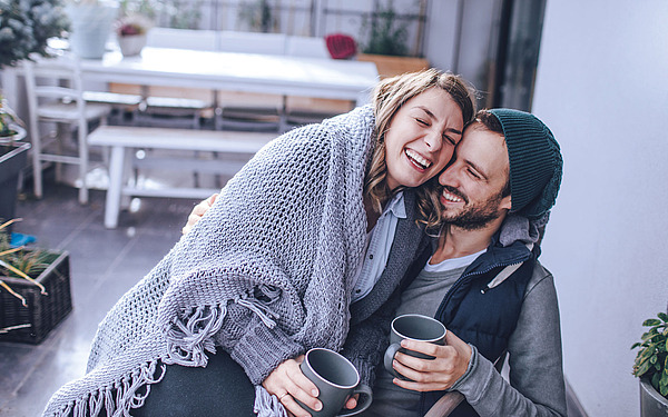 Junges Pärchen sitzt arm in arm mit Kaffeetasse auf dem Balkon