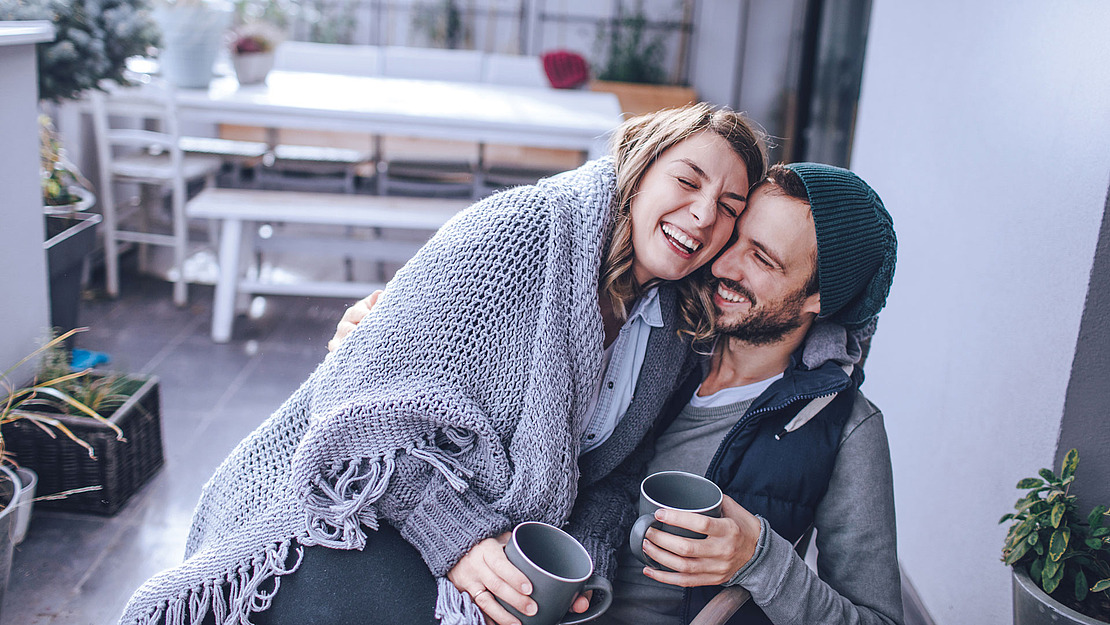 Junges Pärchen sitzt arm in arm mit Kaffeetasse auf dem Balkon