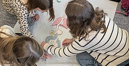 Projekt Kindermedizintag Drei Kinder sitzen auf dem Boden und zeichnen und malen innere Organe auf ein großes Blatt Papier, auf dem der Umriss eines menschlichen Körpers abgebildet ist. Während sie gemeinsam daran arbeiten, sind bunte Filzstifte und ausgeschnittene Organe zu sehen.