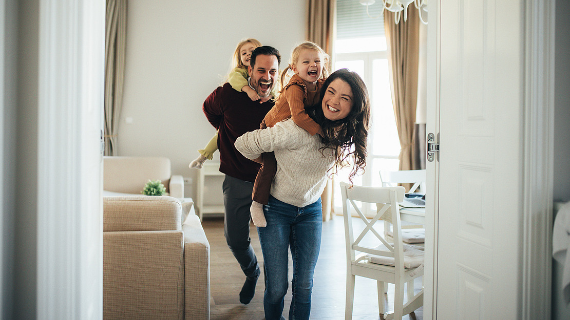 Eine fröhliche vierköpfige Familie spielt im Haus; eine Frau trägt ein kleines Mädchen auf dem Rücken, während ein Mann ein weiteres Kind trägt. Alle lächeln und lachen in einem hellen, gemütlichen Wohnzimmer.