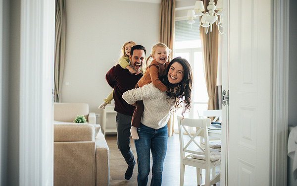 Eine fröhliche vierköpfige Familie spielt im Haus; eine Frau trägt ein kleines Mädchen auf dem Rücken, während ein Mann ein weiteres Kind trägt. Alle lächeln und lachen in einem hellen, gemütlichen Wohnzimmer.
