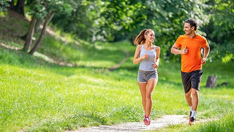 Ein Mann und eine Frau joggen gemeinsam auf einem Kiesweg durch einen grünen Park und lächeln sich dabei an. Sie tragen sportliche Kleidung und die Sonne scheint durch die Bäume um sie herum.