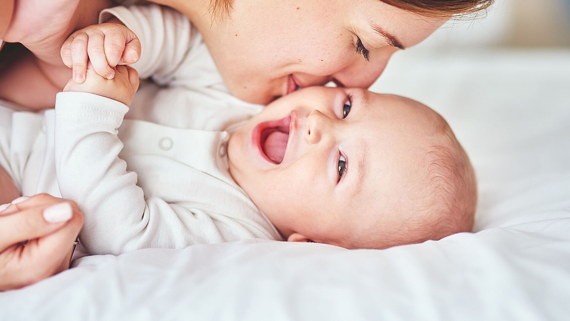 Eine Frau küsst liebevoll ein lächelndes Baby auf die Wange, während beide in einem weißen Bett liegen und helle Kleidung tragen. Das Baby sieht glücklich und fröhlich aus.