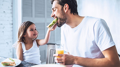 Ein junges Mädchen füttert einen lächelnden erwachsenen Mann mit einem Sandwich und hält ein Glas Orangensaft in der Hand. Sie sitzen zusammen an einem Tisch und tragen beide weiße Hemden.