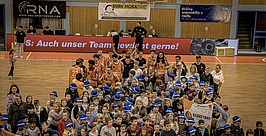 DBB-Grundschulwoche Eine große Gruppe von Kindern posiert mit Basketballspielern auf einem Hallenplatz. Viele Kinder tragen blaue Mützen und halten Basketballartikel in der Hand. Die Spieler tragen orangefarbene Trikots. Im Hintergrund sind Banner und Werbung zu sehen.