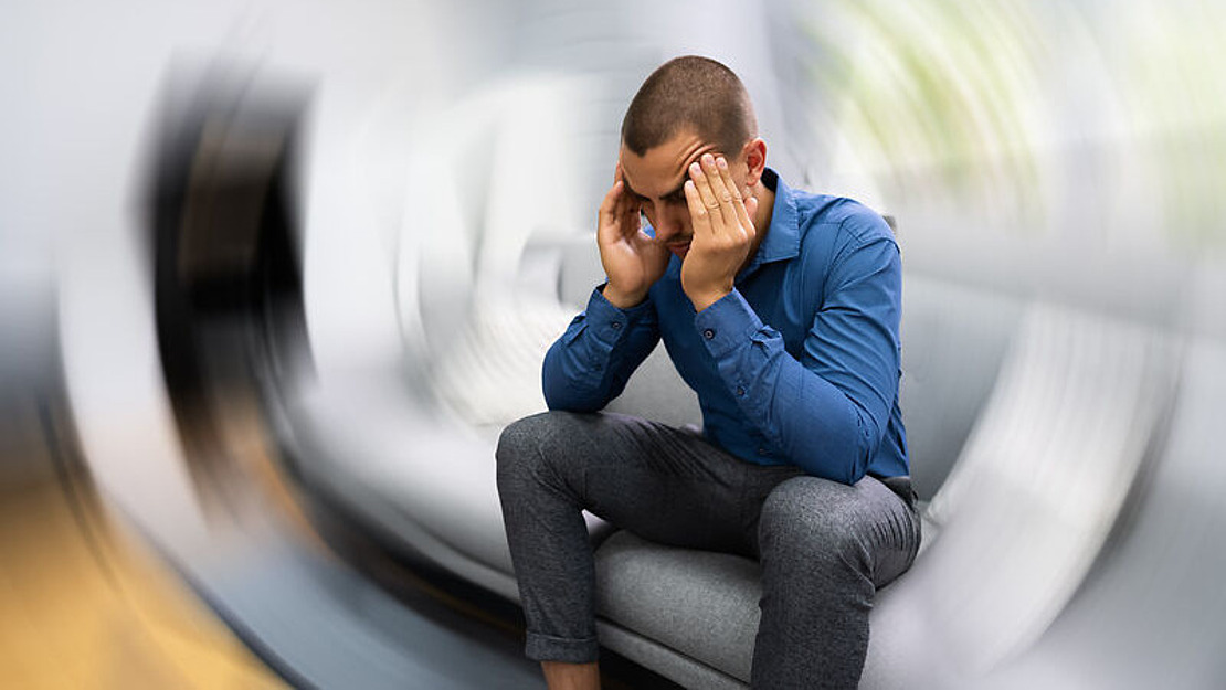 Ein Mann sitzt auf einer Couch, hat die Ellbogen auf die Knie gestützt und die Hände auf die Schläfen gelegt, er wirkt gestresst oder hat Schmerzen. Das Bild hat an den Rändern einen verschwommenen, wirbelnden Effekt, der Schwindel oder Verwirrung suggeriert.