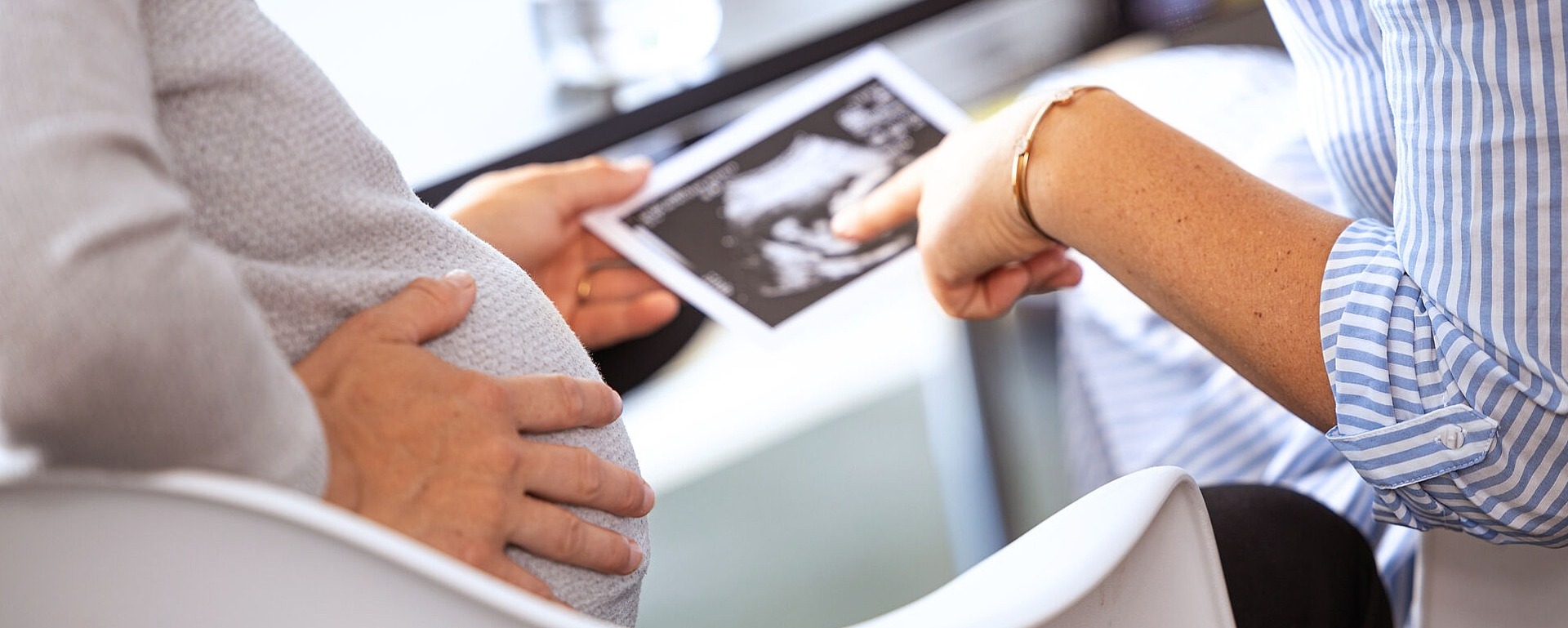 Eine schwangere Person sitzt da und hält ein Ultraschallbild in der Hand, während eine andere Person in einem gestreiften Hemd darauf zeigt. Sie sitzen an einem Tisch, im Hintergrund sind eine teilweise sichtbare Flasche und ein Glas zu sehen.
