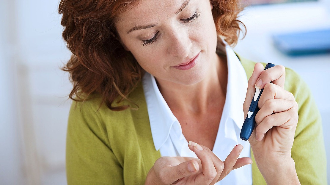 Eine Frau mit roten Locken und grüner Strickjacke benutzt ein Blutzuckermessgerät, um ihren Blutzucker durch Stechen in den Finger zu messen.