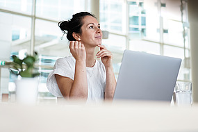 Eine Frau sitzt an einem Schreibtisch mit einem Laptop, blickt auf und lächelt nachdenklich. Sie trägt ein weißes Hemd und befindet sich in einem hellen, modernen Büroraum mit großen Fenstern. Eine Pflanze und ein Glas Wasser stehen auf dem Schreibtisch.
