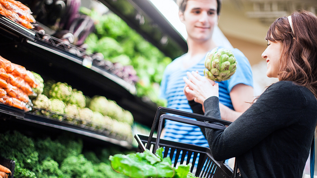 Eine Frau, die einen mit Salat gefüllten Einkaufskorb in der Hand hält, lächelt einen Mann an, während sie in der Gemüseabteilung eines Lebensmittelgeschäfts frisches Gemüse, darunter Artischocken, einkaufen.