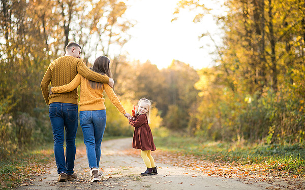 Eine dreiköpfige Familie geht im Herbst einen von Bäumen gesäumten Weg entlang. Die Eltern, die gelbe Pullover tragen, gehen Arm in Arm, während ihre kleine Tochter in einem kastanienbraunen Kleid in die Kamera lächelt und die Hand ihrer Mutter hält.