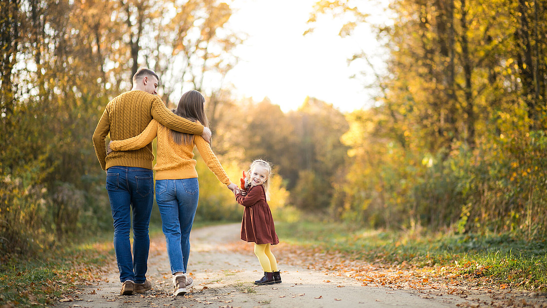 Eine dreiköpfige Familie geht im Herbst einen von Bäumen gesäumten Weg entlang. Die Eltern, die gelbe Pullover tragen, gehen Arm in Arm, während ihre kleine Tochter in einem kastanienbraunen Kleid in die Kamera lächelt und die Hand ihrer Mutter hält.