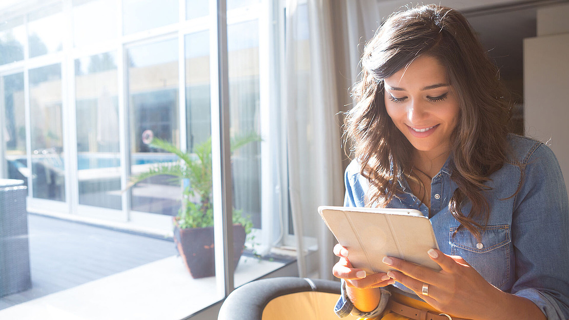 Eine Frau mit langen braunen Haaren und einem Jeanshemd sitzt drinnen an einem hellen Fenster und lächelt, während sie ein Tablet benutzt. Sonnenlicht strömt herein und beleuchtet sie und eine Topfpflanze in der Nähe.