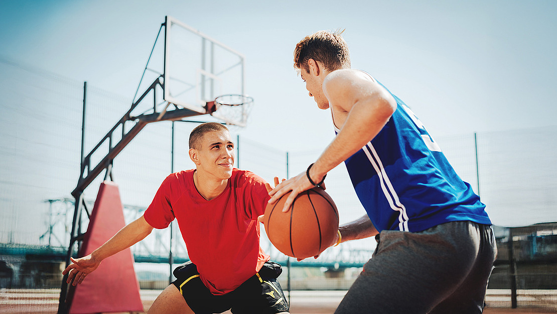 Zwei junge Männer spielen im Freien Basketball. Einer im roten Hemd verteidigt, während der andere im blauen Trikot den Ball dribbelt. Im Hintergrund ist ein Basketballkorb zu sehen, und der Himmel ist klar.