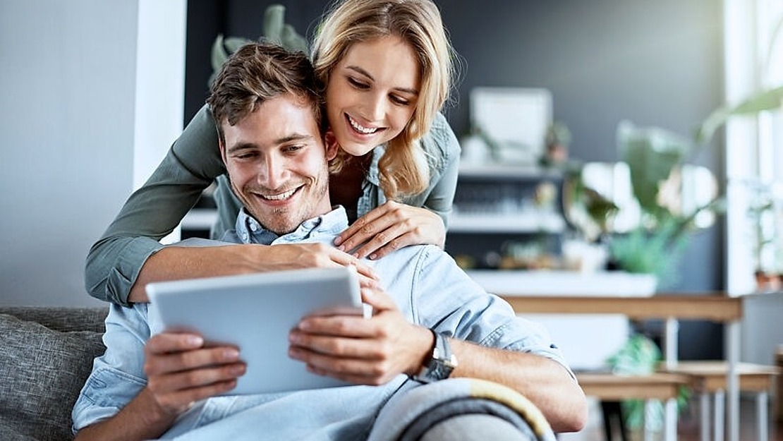 Ein lächelndes Paar sitzt auf einer Couch; der Mann hält ein Tablet, während die Frau ihn von hinten umarmt. Beide wirken glücklich und entspannt in einem hellen, modernen Wohnzimmer.