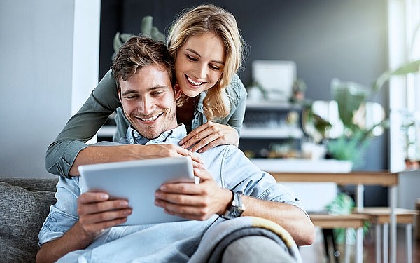 Ein lächelndes Paar sitzt auf einer Couch; der Mann hält ein Tablet, während die Frau ihn von hinten umarmt. Beide wirken glücklich und entspannt in einem hellen, modernen Wohnzimmer.