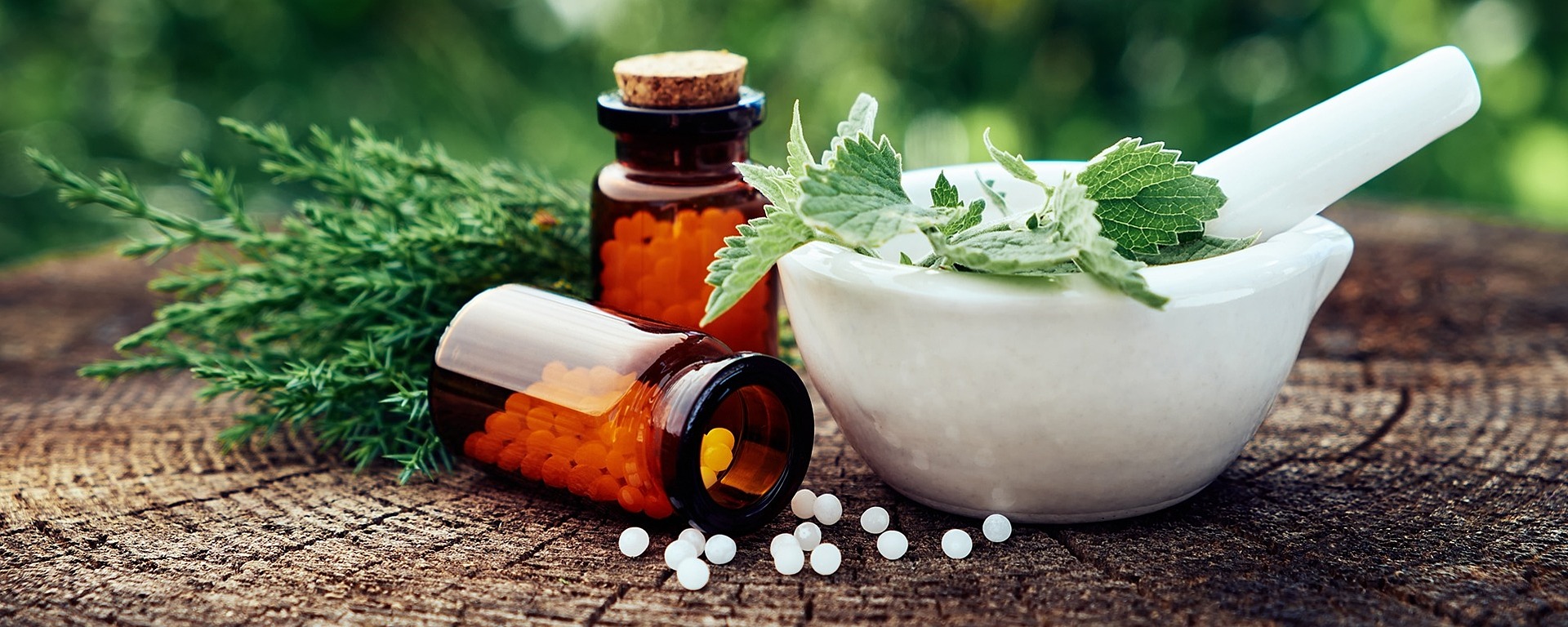 Flaschen mit homöopathischen Arzneimitteln und ein weißer Mörser mit frischen Kräutern liegen auf einer Holzoberfläche mit einem verschwommenen grünen Naturhintergrund.