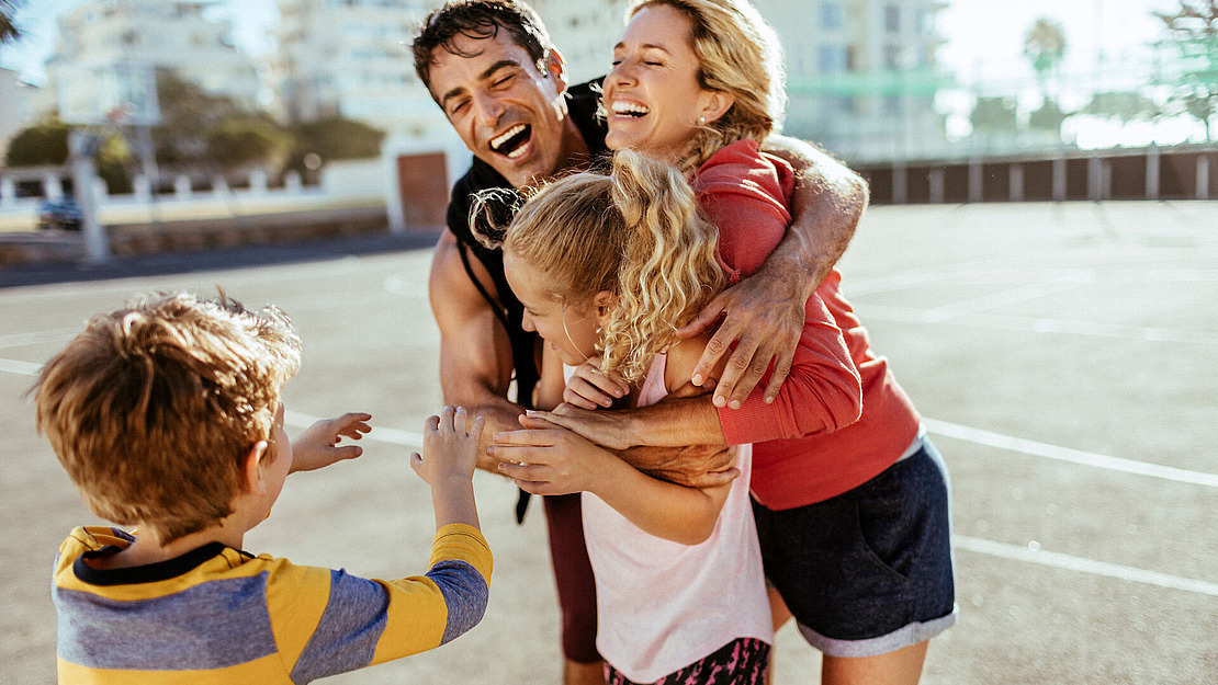 Eine fröhliche vierköpfige Familie, darunter zwei Kinder, umarmt sich und lacht gemeinsam an einem sonnigen Tag auf einem Basketballplatz im Freien.