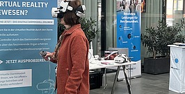 Gesundheitstag bei der vivida bkk in Schwenningen Eine Frau, die ein VR-Headset trägt und einen Controller in der Hand hält, steht vor einem Informationsbanner über ein Virtual-Reality-Modell des Dickdarms.