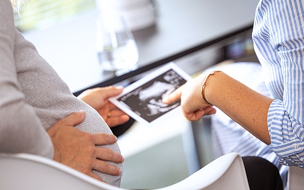 Eine schwangere Person hält ihren Bauch, während eine andere Person in einem gestreiften Hemd auf ein Ultraschallbild zeigt, beide sitzen an einem Tisch mit einem Glas Wasser und Papieren im Hintergrund.