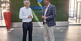 Besuch in der Zentrale der vivida bkk in Villingen-Schwenningen Zwei Männer stehen in einer modernen Lobby mit einer grünen Pflanzenwand, auf der das Wort „vivida“ zu lesen ist, und unterhalten sich. Der eine trägt ein weißes Hemd und eine dunkle Hose, der andere eine karierte Jacke, ein rosa Hemd und eine Khakihose.