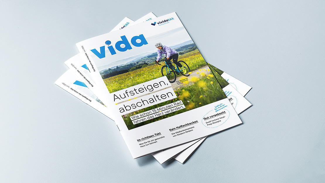 Ein Stapel Zeitschriften mit dem Titel "vida" und dem Titelbild einer Person, die mit dem Fahrrad durch ein Feld mit gelben Blumen unter blauem Himmel fährt. Die Hauptüberschrift lautet: "Aufsteigen, abschalten.