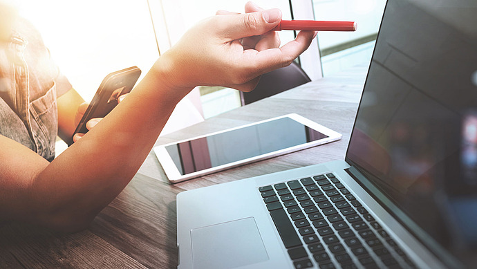 Eine Person hält einen roten Stift und ein Smartphone in der Hand und arbeitet an einem Schreibtisch mit einem geöffneten Laptop und einem Tablet. Im Hintergrund ist ein heller, sonnendurchfluteter Raum mit großen Fenstern zu sehen.