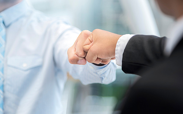 Zwei Menschen in Geschäftskleidung geben sich einen Faustschlag, der Teamwork oder Einigkeit symbolisiert. Der Hintergrund ist unscharf und lenkt den Blick auf die Hände und die Geschäftskleidung.