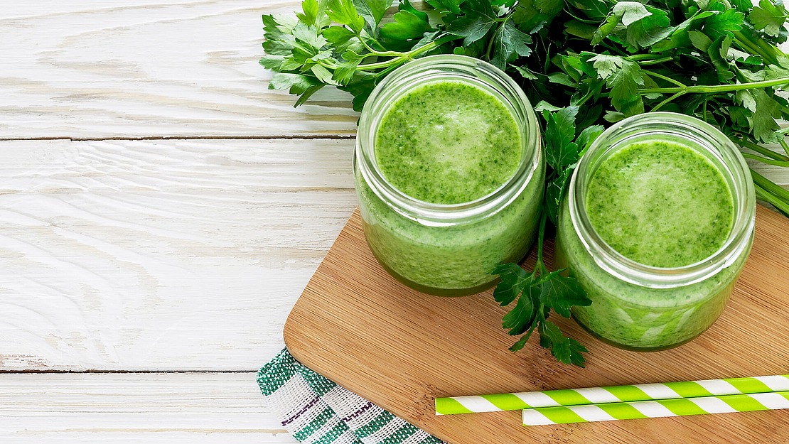 Zwei Gläser mit grünem Smoothie stehen auf einem Holzbrett, umgeben von Blattgemüse. Daneben liegen ein paar grün-weiß gestreifte Strohhalme. Der Hintergrund ist eine helle Holzoberfläche mit einem grün-weiß gestreiften Tuch.