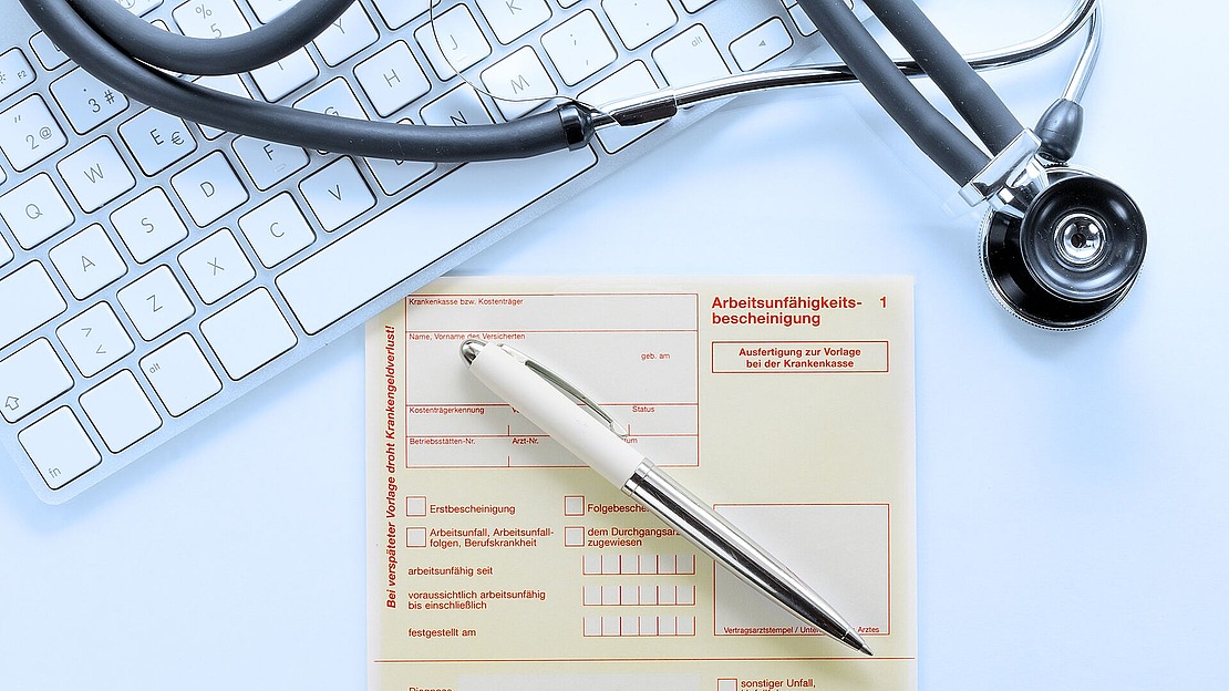 Ein Stethoskop, eine Computertastatur und ein Stift liegen auf einem deutschen ärztlichen Bescheinigungsformular mit dem Titel "Arbeitsunfähigkeitsbescheinigung".