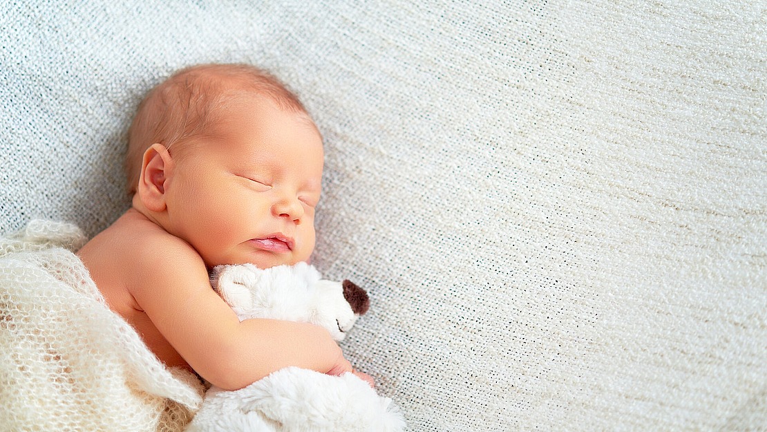 Ein neugeborenes Baby schläft friedlich auf einer weichen, hellen Decke und kuschelt mit einem kleinen weißen Plüschtier. Das Baby ist teilweise in eine kuschelige Strickdecke eingewickelt, während sanftes natürliches Licht die Szene beleuchtet.