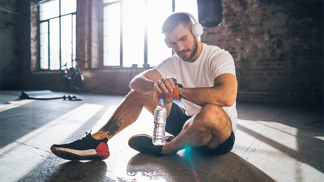 Ein Mann in Sportkleidung sitzt auf dem Boden einer Turnhalle, hält eine Wasserflasche in der Hand und schaut auf seine Smartwatch. Er trägt Kopfhörer und ruht sich nach dem Training aus, während Sonnenlicht durch die großen Fenster hinter ihm strömt.
