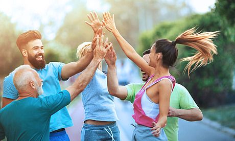 Eine Gruppe von fünf Personen unterschiedlichen Alters, in legerer Sportkleidung, geben sich im Freien fröhlich High Fives. Sie wirken voller Energie und glücklich, umgeben von viel Grün an einem sonnigen Tag.