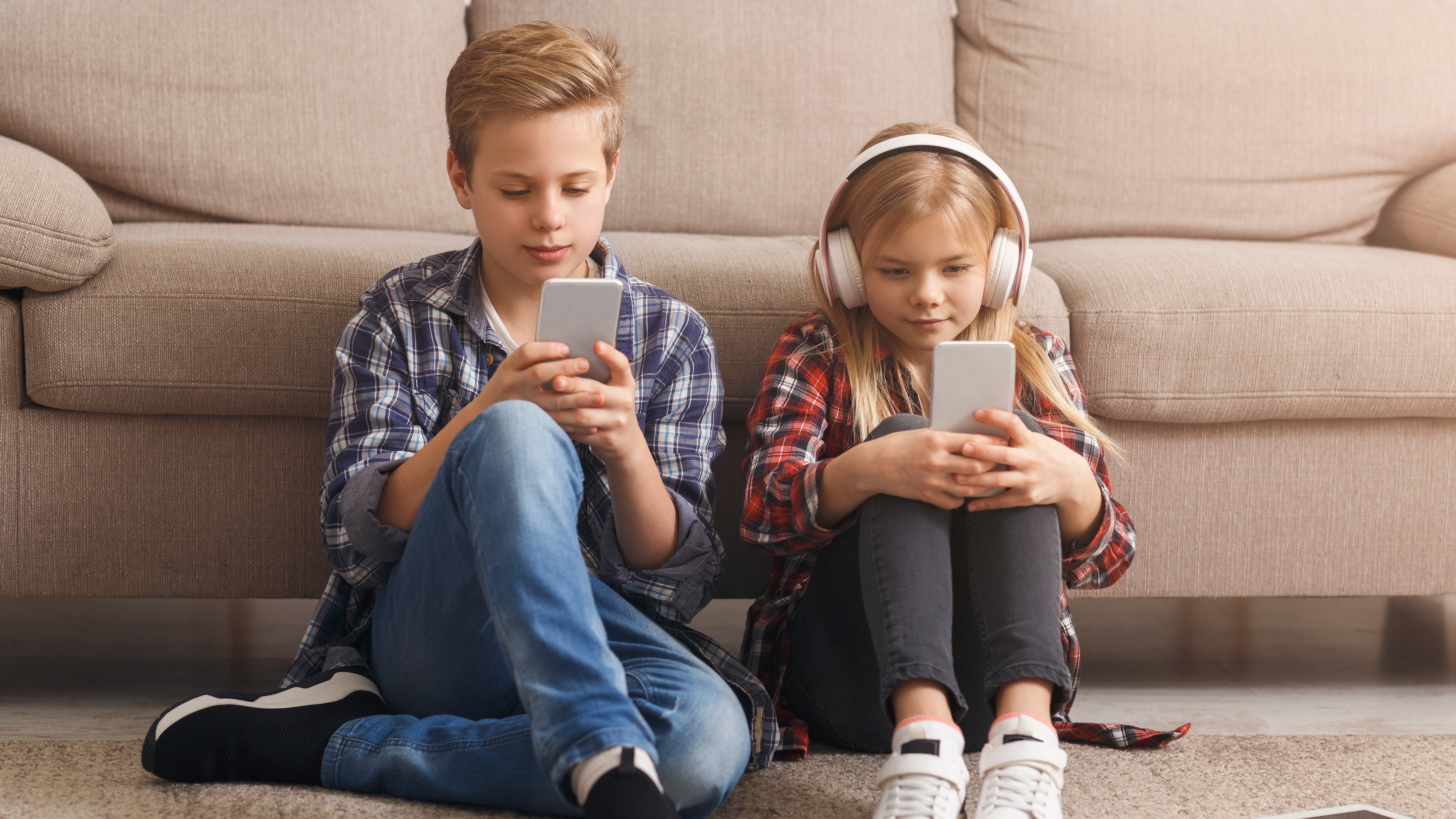 Zwei Kinder sitzen auf einem Teppich vor einem Sofa und konzentrieren sich auf ihre Smartphones. Der Junge trägt ein kariertes Hemd und Jeans, das Mädchen Kopfhörer und ein kariertes Hemd. Bücher und ein Tablet liegen daneben auf dem Boden.