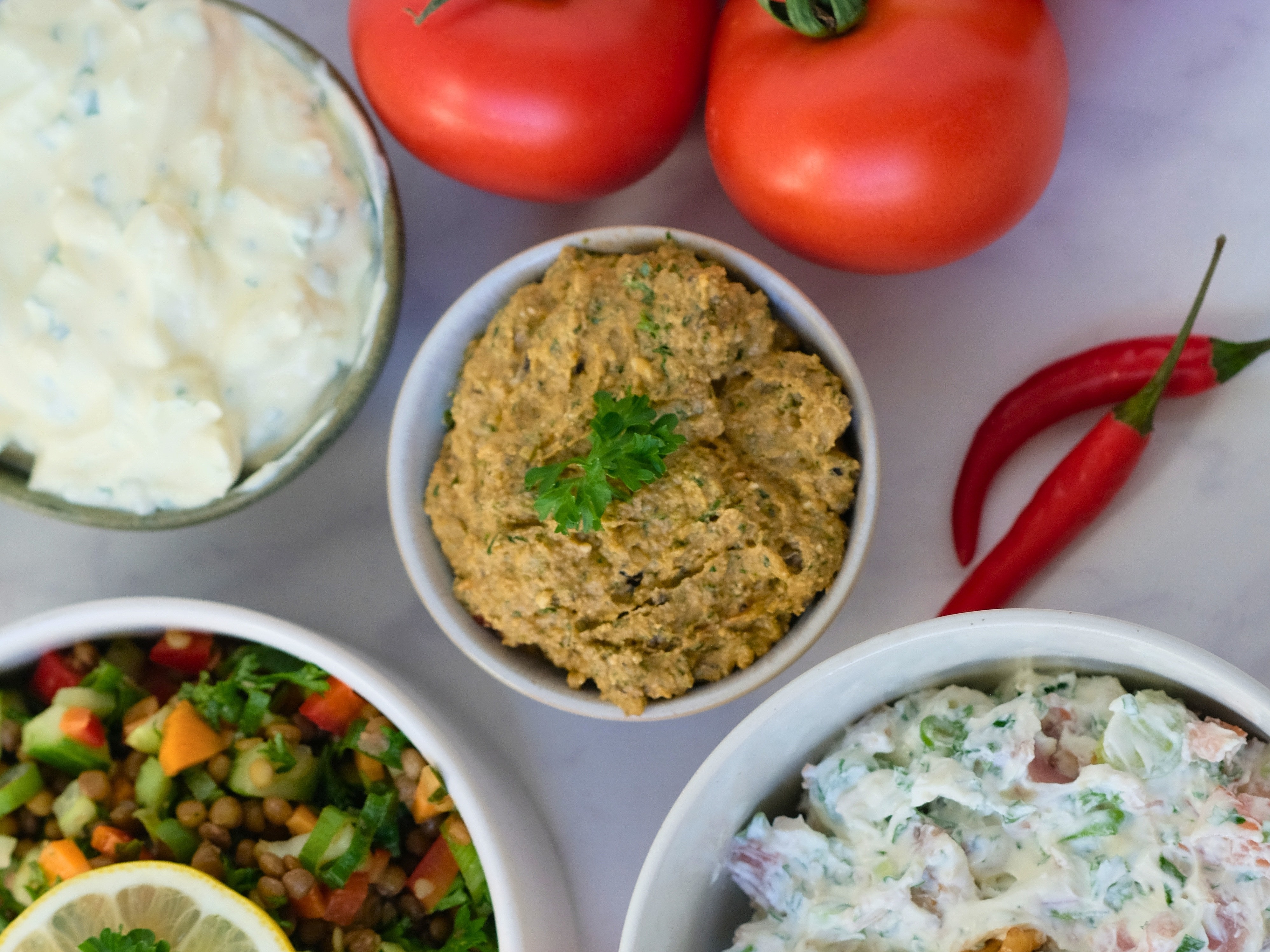 Draufsicht auf vier mit verschiedenen Dips und Salaten gefüllte Schalen, umgeben von frischen Tomaten, roten Chilischoten, einer roten Paprika und Walnüssen auf einer hellen Fläche.