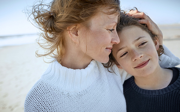 Eine Frau legt ihre Hand sanft auf den Kopf eines Kindes, während beide lächeln. Sie stehen dicht beieinander an einem Strand, im Hintergrund ist das Meer zu sehen. Die Frau trägt einen weißen Pullover und das Kind einen dunklen.