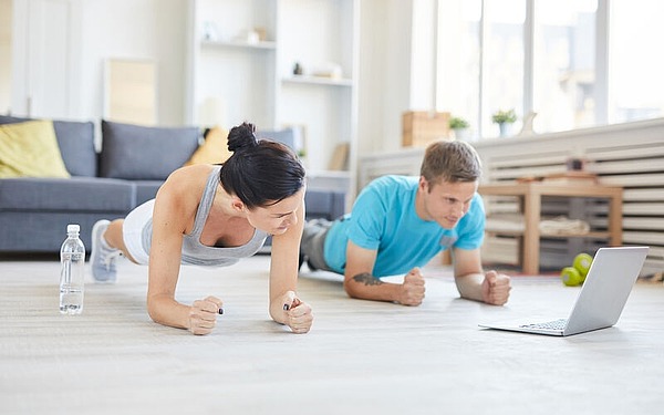 Eine Frau und ein Mann trainieren gemeinsam zu Hause. Sie machen Planken auf einem hellen Boden und schauen sich dabei ein Workout auf einem Laptop an. Eine Wasserflasche und Hanteln stehen in der Nähe, im Hintergrund ein Sofa und Regale.