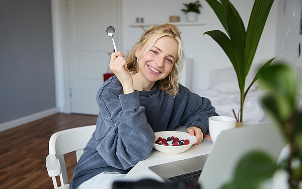 Eine lächelnde Frau sitzt an einem Tisch in einem hellen Raum, in der Hand einen Löffel und eine Schale mit Obst und Joghurt. Eine Kaffeetasse, ein Laptop und eine Grünpflanze stehen ebenfalls auf dem Tisch. Sie wirkt entspannt und fröhlich.