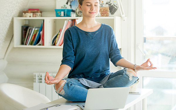 Eine Person sitzt im Schneidersitz auf einem Schreibtisch vor einem Laptop, die Augen sind geschlossen und die Hände ruhen in Meditationshaltung auf den Knien. Im Hintergrund sind Bücher und Regale zu sehen, und das Sonnenlicht erfüllt den Raum.