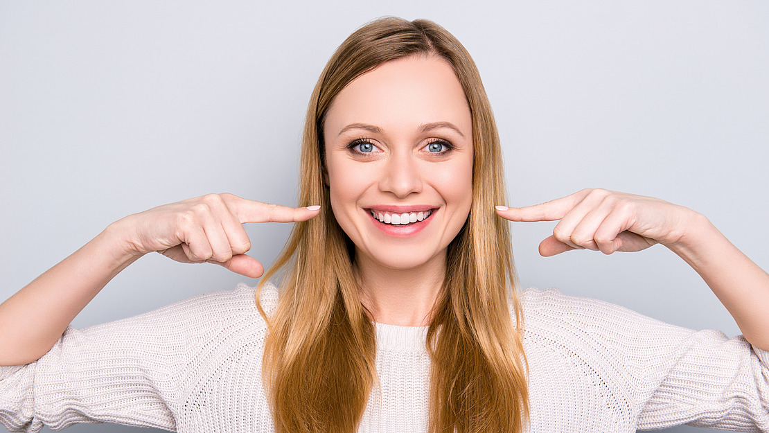 Eine lächelnde Frau mit langen blonden Haaren zeigt mit beiden Zeigefingern auf ihre Zähne. Sie trägt einen hellen Pullover und steht vor einem schlichten, hellen Hintergrund.