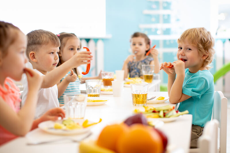 Eine Gruppe kleiner Kinder, die an einem Tisch sitzen, essen und Saft trinken, mit Tellern voller gesunder Mahlzeiten und Obst in einem hellen, freundlichen Raum.