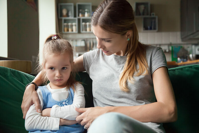 Ein junges Mädchen sitzt mit verschränkten Armen auf einer Couch und sieht verstört aus. Neben ihr hat eine Frau den Arm um das Mädchen gelegt und scheint sanft mit ihr zu sprechen, um sie zu trösten oder ihr etwas zu erklären.