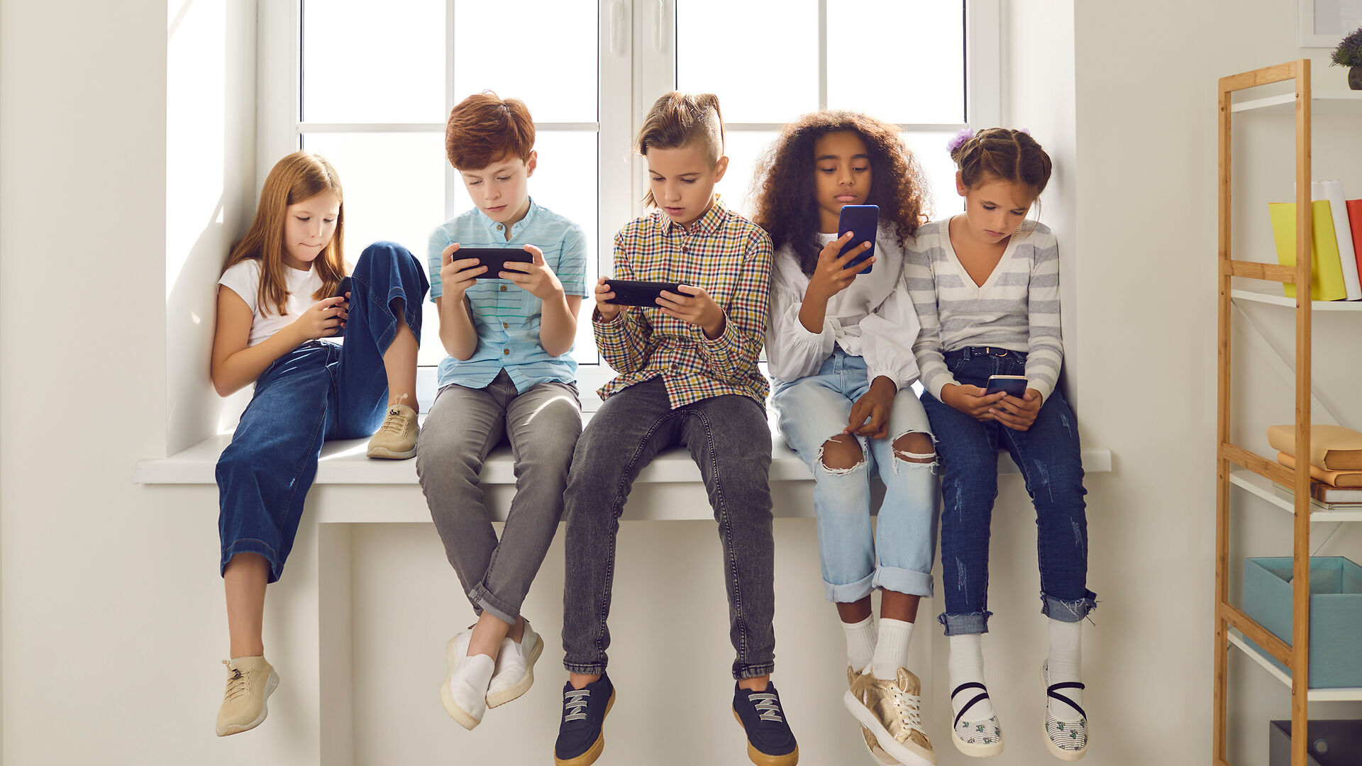 Fünf Kinder sitzen auf einer Fensterbank, jedes ist auf ein Smartphone oder ein anderes mobiles Gerät konzentriert. Sonnenlicht fällt durch das Fenster hinter ihnen. Auf der rechten Seite sind Regale und Bücher zu sehen.