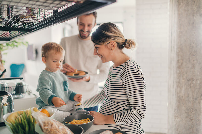 Eine lächelnde Familie in einer Küche: eine schwangere Frau bereitet das Essen zu, während ein kleines Kind auf dem Tresen sitzt und ein Mann in der Nähe steht und einen Teller hält. Sie scheinen das Frühstück gemeinsam zu genießen.