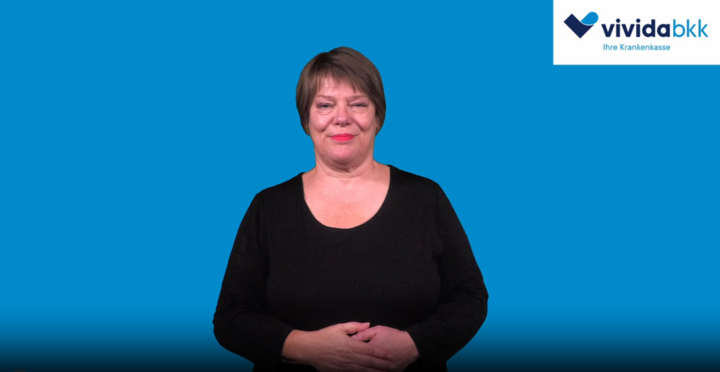 Eine Frau mit kurzen braunen Haaren, die ein schwarzes Oberteil trägt, gestikuliert mit ihrer rechten Hand vor einem blauen Hintergrund. In der rechten oberen Ecke erscheint das Logo von vivida bkk.